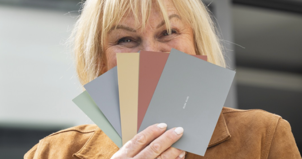Nadine Rapp, décoratrice d'intérieur à Mulhouse, tenant un nuancier de teintes douces — gris bleuté, terracotta, vert sauge — devant son visage, en extérieur